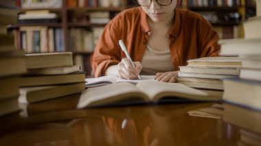 Gözlüklü kız öğrenci not alıyor, üniversite kütüphanesinde kitap okuyor.