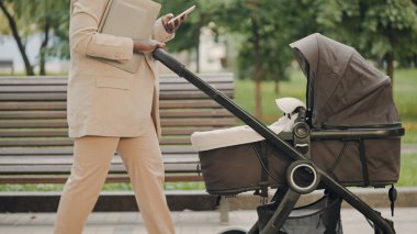 Resmi takım elbiseli bir kadın akıllı telefondan e-posta yolluyor, açık havada bebek arabasıyla yürüyor.