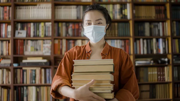 Positive asian student in mask holding stack of books, education during pandemic