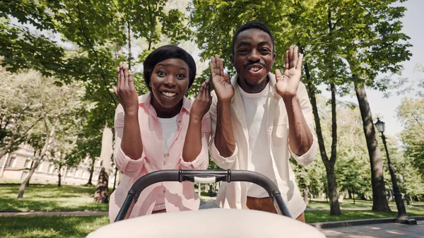 Funny black parents playing peek-a-boo with a baby in stroller, making child laugh