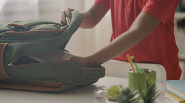 Çocuğun elleri sırt çantasına kitap koyuyor, çocuk yeni okul gününe hazırlanıyor.