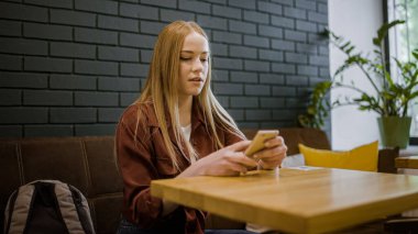 Genç sarışın kadın kahve masasının arkasında oturuyor, akıllı telefon uygulaması kullanıyor.