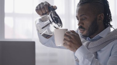 Uykulu Afrikalı Amerikalı adam dizüstü bilgisayarla çalışıyor, kahvesini bir fincana dolduruyor.