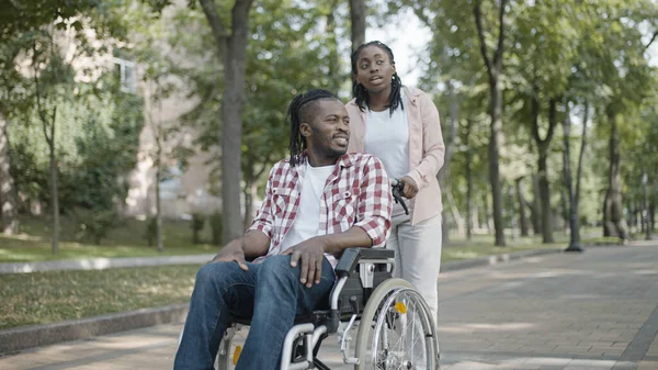 Black nurse pushing wheelchair with male patient in hospital park, recovery