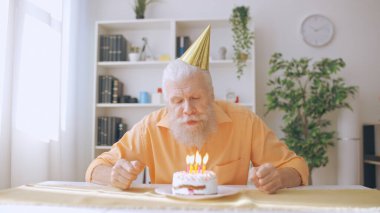Mature man blows out candles on his birthday cake, marking his birthday alone during the coronavirus lockdown