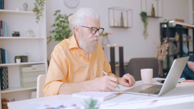 Focused senior man works from home, browsing the Internet and jotting down ideas