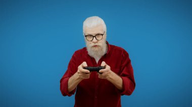 Senior man presses buttons on a joystick against a blue background, embracing his inner video game geek