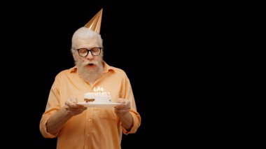 Smiling senior man blows out candles on a birthday cake against a black background