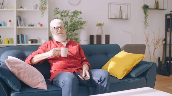 A contemporary grandpa relaxes by watching TV and drinking tea at home, comfortably seated on a sofa