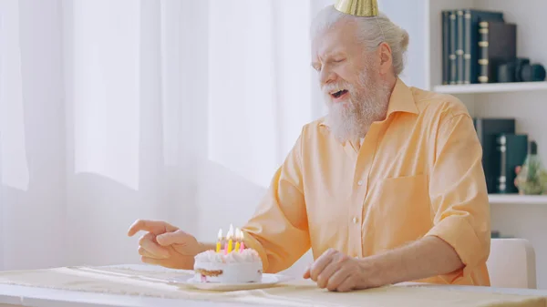 A gray-haired man sits in a birthday hat, about to blow out the candles on a cake, celebrating his birthday amid lockdown