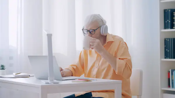 A gray-haired man watches videos on his laptop and enjoys a warm beverage, spending quality time at home