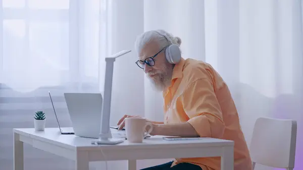 Retired man in a headset works or plays video games on his laptop, indulging in his hobby at home