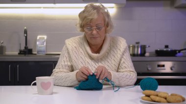 Sarışın, yaşlı bir kadın masada örgü örüyor, evde emeklilik ve rahatlığın tadını çıkarıyor.