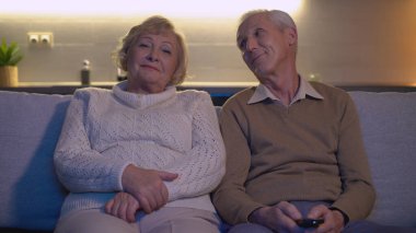 Yaşlı bir adam karısına sevgiyle bakıyor, çiftler birlikte televizyon seyrediyor.