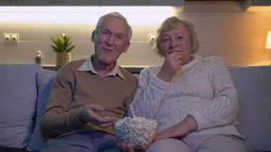 Emekli bir karı koca kanepede komik bir film izleyip birlikte patlamış mısır yiyorlar.
