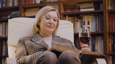 50 'li yaşlardaki zengin bir kadın kitap okuyor, şarap içiyor, kütüphanede dinleniyor.