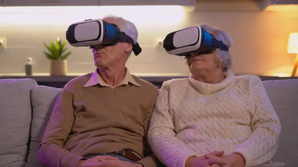A mature couple is trying modern technologies, sitting on the sofa with virtual reality headsets