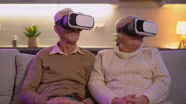 The couple is visiting an exhibition online, sitting on the sofa with 3D headsets, experiencing future innovation