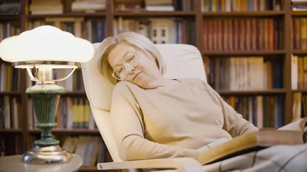 Senior woman falling asleep while reading a book in her library, late evening
