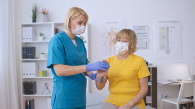 Koruyucu yüz maskesi takmış özenli bir doktor yaşlı bir kadının elini artrit tedavisi için muayene eder.