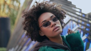 Siyah gözlüklü, kıvırcık saçlı, kendine güvenen kadın kameraya bakıyor.