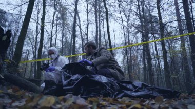 Erkek kriminalist, kurbanın cesedini görmek için plastik bir torba açıyor. Ormandaki suç mahallinde çalışıyor.