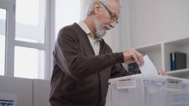 Vatandaşlık görevini yerine getiren yaşlı bir adam oy pusulasını bir konteynıra atıyor.