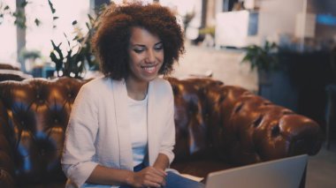 Kafedeki genç kadın dizüstü bilgisayarda çalışıyor, uzaktan görüşme yapıyor, rahat bir kafe ortamı yaratıyor.