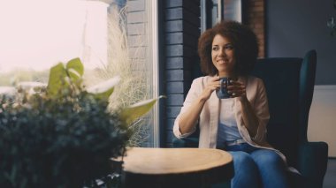 Gülümseyen siyah kadın kahve içiyor ve sıcak kahve dükkanında pencereden dışarı bakıyor.
