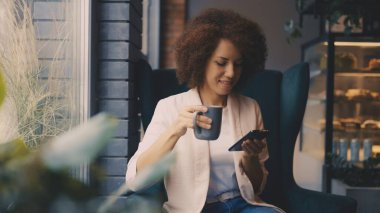 Neşeli siyah kadın kahve içip telefon kullanıyor, sabahları eğleniyor.