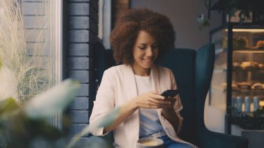 Neşeli siyah kadın kahvesini yudumluyor ve rahat kafe ortamında telefonunu kontrol ediyor.