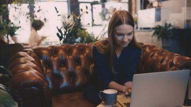 Gülümseyen kadın içkisini yudumluyor ve kafeteryada dizüstü bilgisayarla çalışıyor.