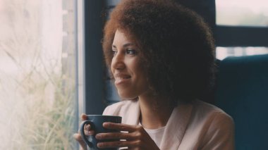 Neşeli kıvırcık saçlı kadın kafede sıcak kahve içiyor, sabah rutinine başlıyor.