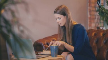Kahvede dizüstü bilgisayarla çalışırken pasta yiyen genç bir kadın, öğle yemeği molası.