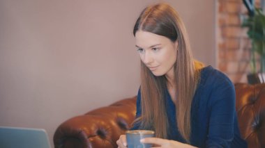 Bir kadın yemek molasında laptopta video izlerken sıcak içecek içiyor.