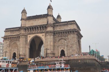 Hindistan 'ın Mumbai' deki ikonik geçidi, görünür kalabalık ve tekneler yakınlarda. Gökyüzü bulutlu, tarihsel atmosfere yeni bir hava katıyor.