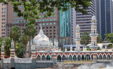 Kuala Lumpur 'daki en eski camilerden biri olan Mescid Jamek Camii. Beyaz kubbeleri ve minareleri modern gökdelenlerin arka planında göze çarpıyor. Cami, ön planda palmiye ağaçları ve bir su özelliği ile çevrilidir..