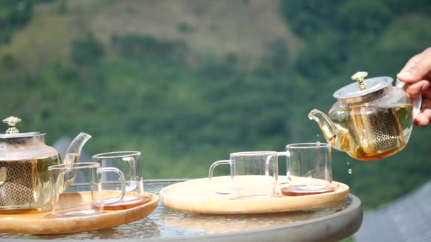 木製のトレイにガラスティーポットからお茶を注ぐ手 ティーポットからお茶を注ぐ手 伝統的な茶器に紅茶を注ぐクロップショット — ストック動画