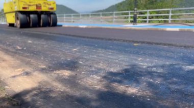 Yol silindiri yeni yerleştirilmiş asfaltı bir yol inşaatına sıkıştırıyor. Sarı patenci yolun pürüzsüz bir bölümünde çalışırken görülüyor, etrafındaki korkuluklar, inşaat mühendisliği...