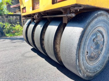 Yeni yerleştirilmiş asfalt, makine gücü ve ağırlığı, yol yapım süreci ve makine detaylarını sıkıştıran ağır yol pateni, yeşillik ve sanayi bölgesinin arka planında yer alıyor.