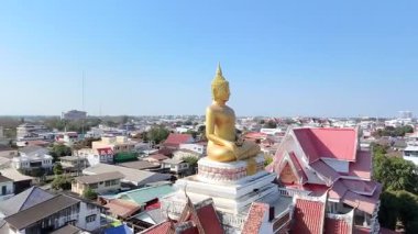 Geleneksel çatısı olan ve açık mavi gökyüzünün altındaki kasaba manzaralı Tayland tapınağındaki büyük altın Buda heykelinin havadan görüntüsü.