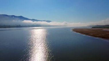 Güneş ışığını yansıtan sakin bir nehrin hava manzarası, sisli dağların manzaralı arkaplanı ve kumlu bir kıyı şeridi. Huzurlu atmosfer, sabahın erken saatlerinde doğanın güzelliğini yakalar.