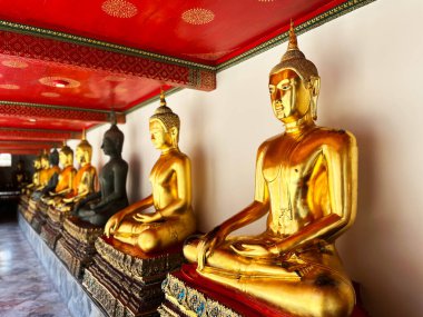 Tayland tapınağının içinde altın Buda heykelleri. Karmaşık kırmızı ve altın tavan süslemeleri var. Heykeller yumuşak doğal ışıkla aydınlatılıyor.