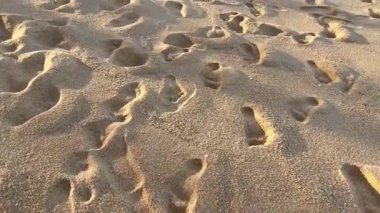 Sahilde yaprak, kumlu bir sahilde ayak izlerinin yakın çekimi, sıcak güneş ışığıyla aydınlatılmış. Kumun dokulu yüzeyi derinlik yaratır, seyahat, yalnızlık, kıyı keşfi hissi uyandırır.