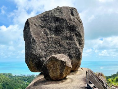 Turkuaz okyanusa bakan bir tepeye tünemiş kaya oluşumu. Engebeli kaya kütleçekimine meydan okuyor gibi görünüyor. Çarpıcı bir doğa simgesi, yeşillik ve dağınık bulutlarla dolu mavi bir gökyüzü yaratıyor.