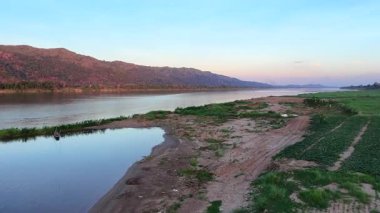 Mekong Nehri, berrak bir gökyüzünün altındaki su, ağaçlar, tepeler, güneş ışığını yansıtan sakin bir nehrin havadan görünüşü, sisli dağların manzaralı arkaplanı.