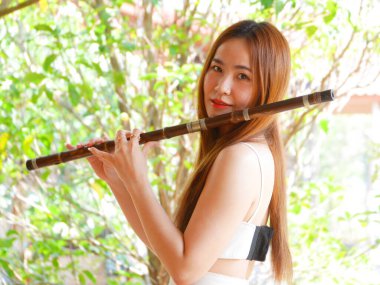 Genç Asyalı bir kadın açık havada bambu flüt çalıyor arka planda yeşil doğa, geleneksel müzik ve kültürel yaşam tarzı kavramı, Asya 'lı genç bir kadın doğada flüt çalıyor.