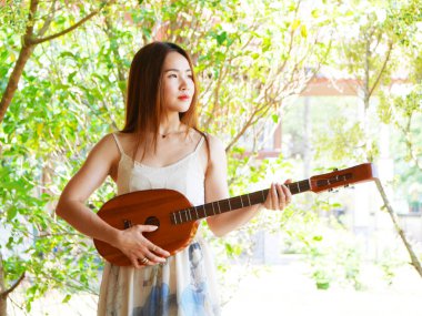 Genç Asyalı bir kadın açık havada akustik bir gitar tutuyor. Müzikal hayat tarzının ve kişisel ifadenin huzurlu anı, gitarlı genç kadın.