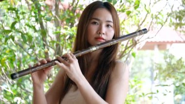 Genç Asyalı bir kadın açık havada bambu flüt çalıyor arka planda yeşil doğa, geleneksel müzik ve kültürel yaşam tarzı kavramı, Asya 'lı genç bir kadın doğada flüt çalıyor.