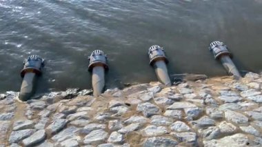 Beş drenaj borusu kayalık bir set boyunca dizilmiş, altın güneş ışığı altında bir göle boşalıyor, su yönetimi altyapısı ve mühendisliği sergiliyor.